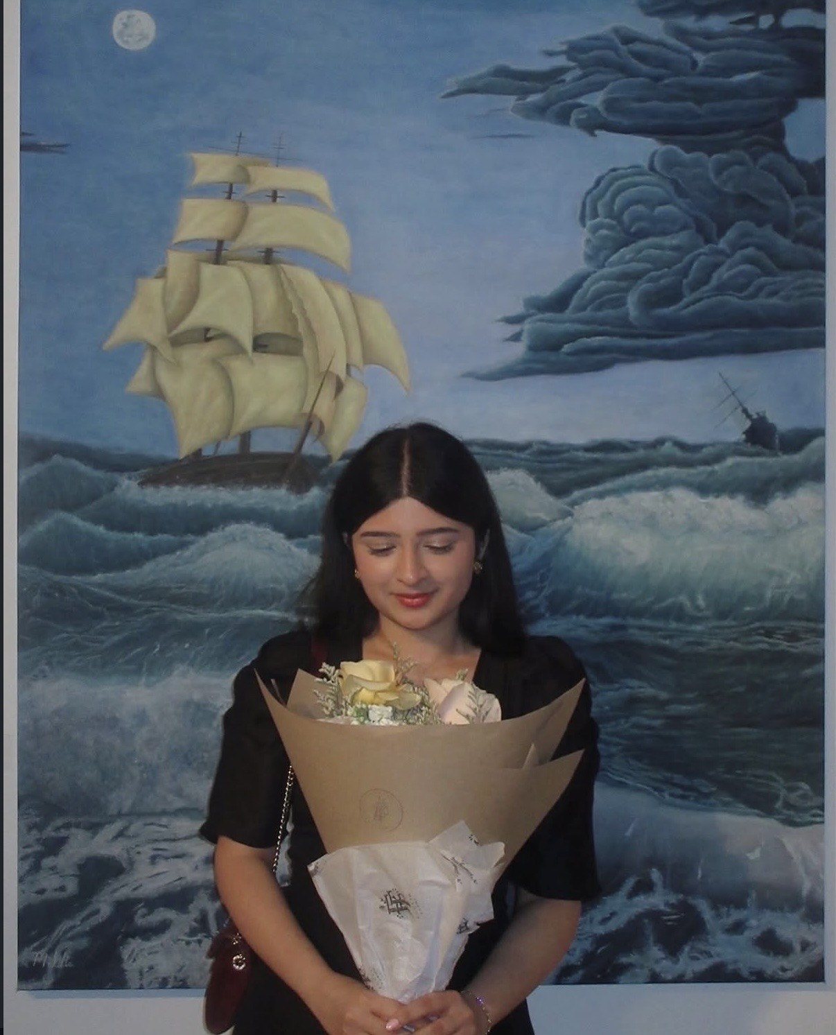 Malika Sharma holding a paper-wrapped bouquet of cream roses, standing before her painting of a tall ship on a moonlit, storm-tossed sea.